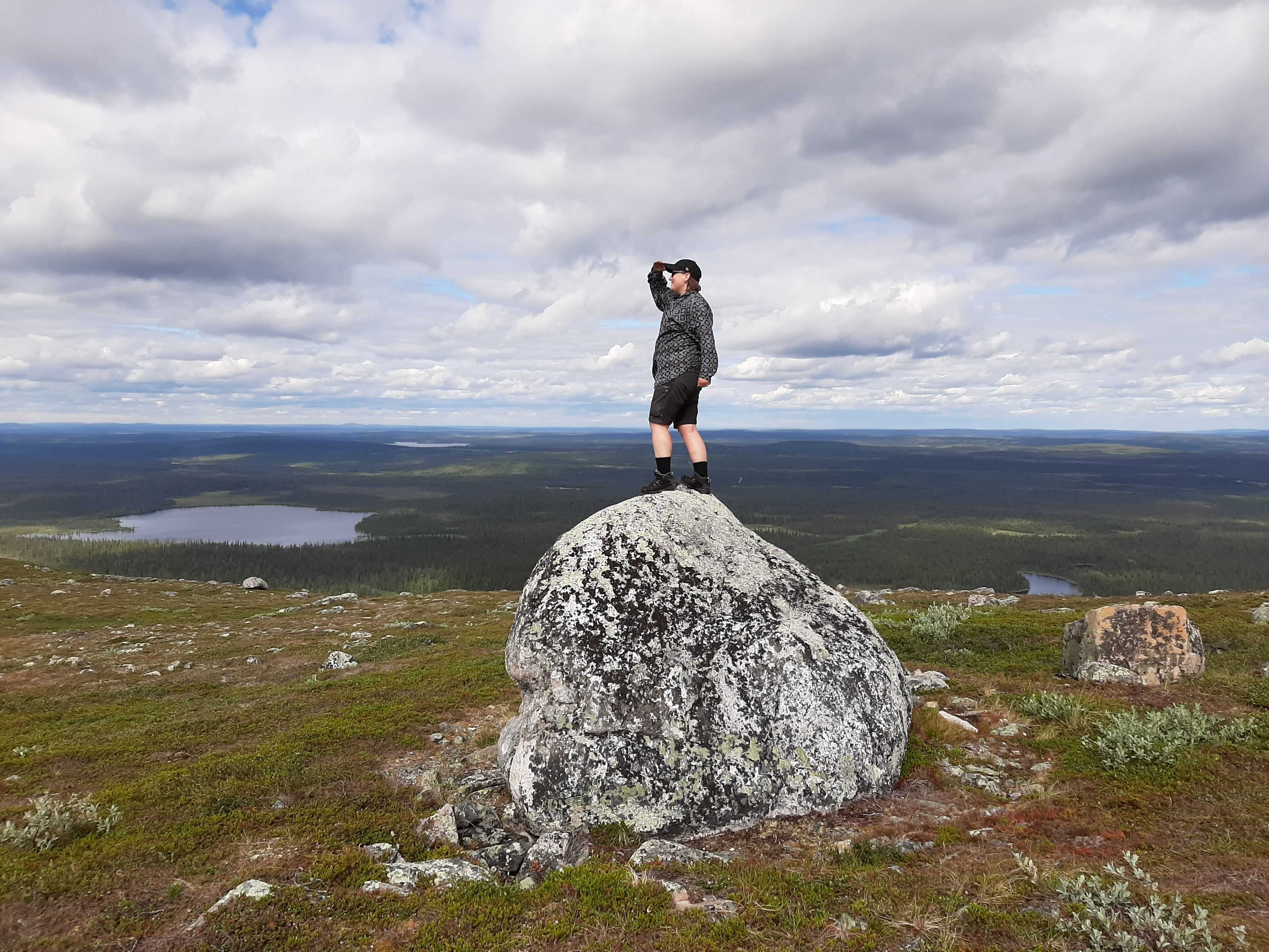 Elisa Raittila tunturimaisemissa kiiveenneenä tähyilemään maisemaa ison siirtolohkareen päältä.